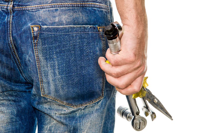 Die Bauhelferversicherung Nahaufnahme einer Person in Jeans, die verschiedene Werkzeuge wie Zange und Schraubenschlüssel in der Hand hält.